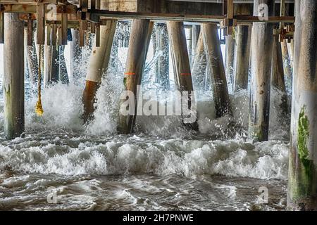 Waves rush in through the pilings of an old pier and result in water splashing energetically. Stock Photo