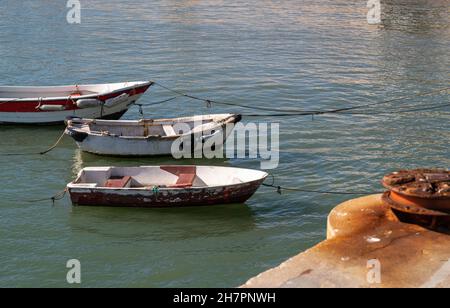 View of three old flaked wooden boats moored to a pier on the right with a part of a rusty bollard on the concrete surface in a defocused foreground, Stock Photo