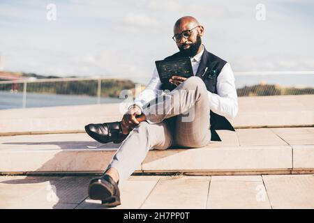 A handsome bald bearded black man entrepreneur in eyeglasses and a formal suit with a vest, is sitting outdoors and interacting with a futuristic tabl Stock Photo