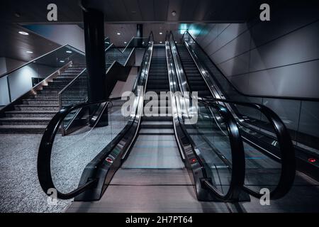 A frontal wide-angle shot of two escalators in a shopping mall or an airport terminal, or a transport station depot, with a staircase on the left; neo Stock Photo