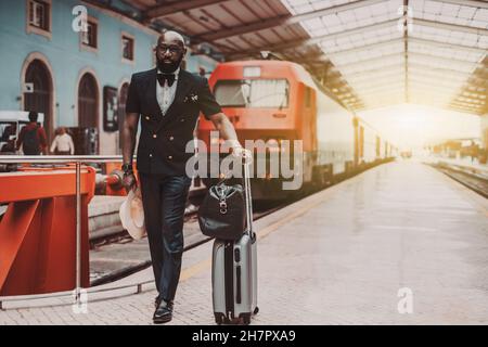 A dapper bearded bald black senior businessman in a fashionable suit with a vest and bowtie is standing on a platform of a railway station depot with Stock Photo