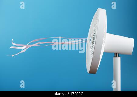 The electric fan is white with pink ribbons fluttering in the wind on a blue background. Free space, minimalistic style. Stock Photo