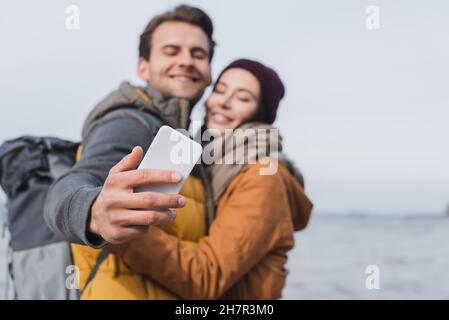 blurred couple in autumn clothes taking selfie on mobile phone while walking near river Stock Photo