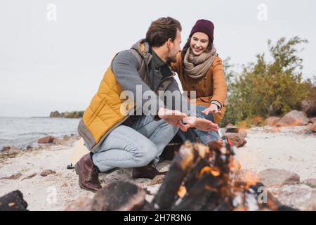 young hikers in autumn clothes warming near blurred bonfire on riverside Stock Photo