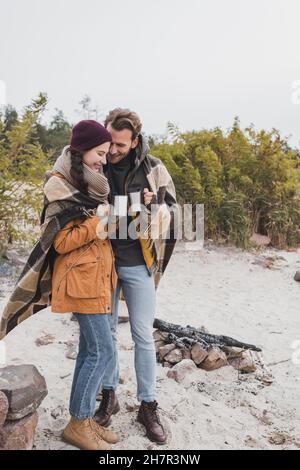 happy couple with thermo cups wrapping in plaid blanket while standing near bonfire Stock Photo