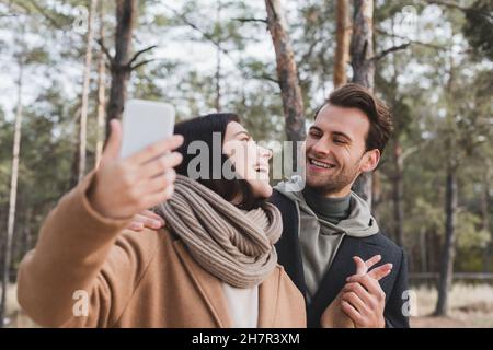 cheerful woman taking selfie on blurred cellphone with boyfriend during walk in forest Stock Photo