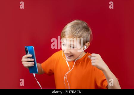 a handsome boy in a yellow T-shirt, who stands on a red background, holds a blue phone in his hand and listens to music on headphones Stock Photo