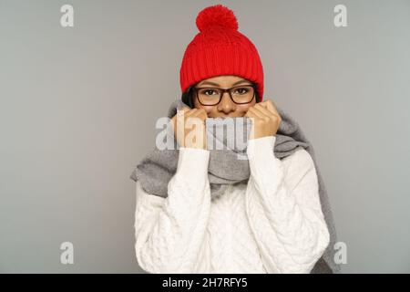 Funny young african woman in sweater, knitted woolen hat hiding smile under warm scarf covering face Stock Photo