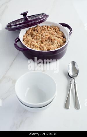 Fresh baked apple crumble in small casserole dish on marble background with bowls and spoons in vertical orientation. Natural stone texture. Stock Photo
