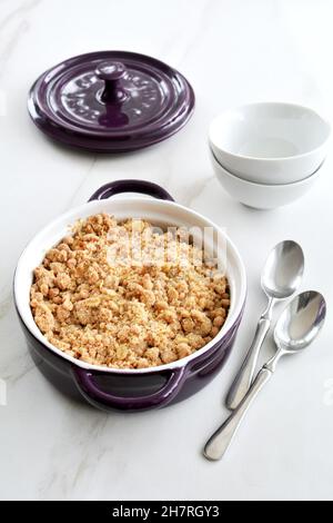 Fresh baked apple crumble in small casserole dish on marble background with bowls and spoons in vertical orientation. Natural stone texture. Stock Photo
