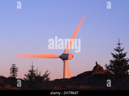 Single wind turbine nacelle and moving blades turned pink by the setting sun behind the summit cairn of an adjacent hill under a clear blue sky. Stock Photo