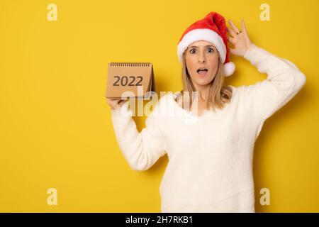 Young amazed woman wearing santa hat showing calendar isolated over yellow background. Stock Photo