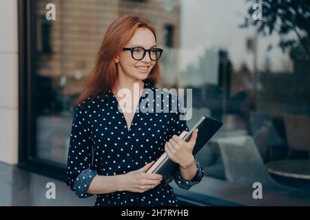 Businesswoman with red hair carries modern device and notepad goes on formal meeting Stock Photo
