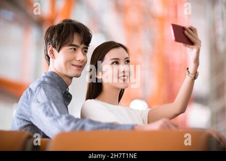 Young couple taking selfies on their mobile phone in an airport lounge Stock Photo