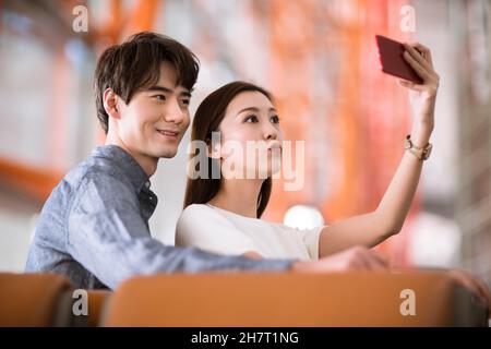 Young couple taking selfies on their mobile phone in an airport lounge Stock Photo