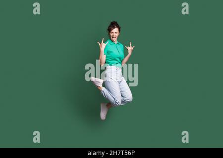 Full size photo of young crazy caucasian woman jumping showing tongue. Rock'n'roll sign. Isolated on green color background Stock Photo