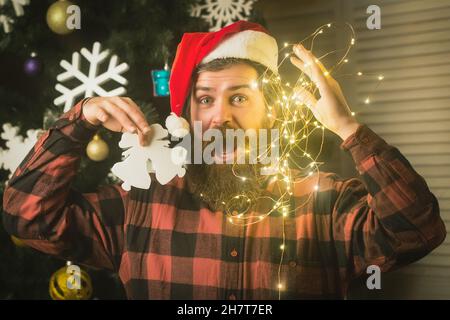 Christmas garland at face of man in hat with beard hold toy, new year holiday and party celebration. Bearded santa face Stock Photo