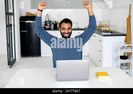 Excited Indian bearded male entrepreneur showing triumph gesture and screaming yes, young hispanic man in casual wear sits at the desk and using laptop indoor, celebrating good offer, finished project Stock Photo