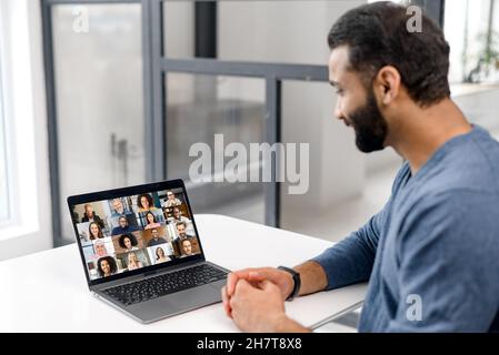View from back above shoulder of Indian guy at the laptop with diverse employees, coworkers on the screen, video call, online meeting. App for video conference with many people together Stock Photo