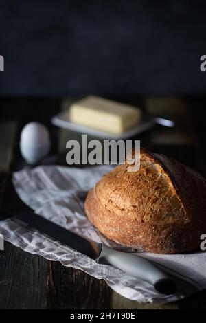 Homemade sourdough wheat bread on a dark background with an egg in a ceramic stand and butter, next to a bread knife. Dark background, a wooden table. Stock Photo