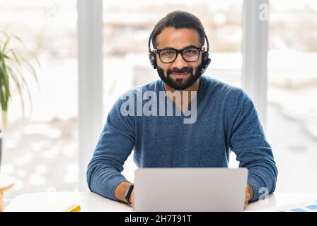 Indian male salesman in a headset is holding the call, ethnic customer service representative sitting in front of a laptop and looking at the camera Stock Photo