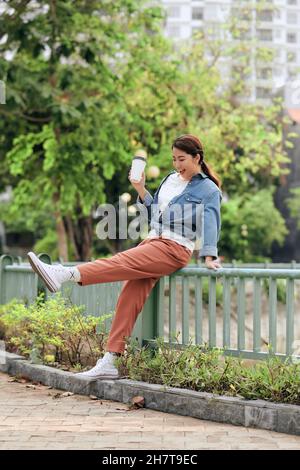 Attractive cheerful trendy girl in stylish denim jacket drinks takeaway coffee or tea outdoors Stock Photo
