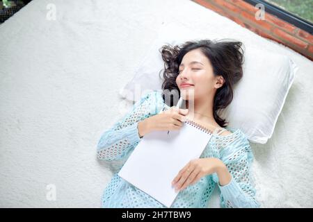 Smart pensive girl lying in bed and making notes in notebook at home Stock Photo