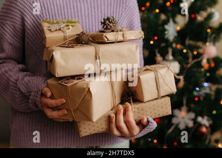 Closeup of woman in violet pullover holding many gift boxes in her hands. Decorated Christmas tree on background. Gift boxes wrapped in craft paper Stock Photo