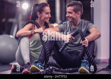 A couple that finished battle ropes workout sit together on a ground inside the gym, looking at each other, smiling. Stock Photo