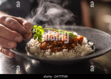Chef decorating vegetarian curry with greens, served over rice on a rustic plate. Stock Photo