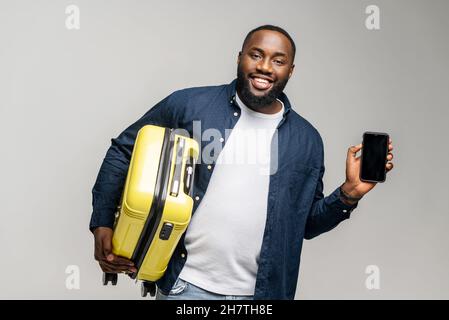 Smiling young African-American guy in casual wear stands with yellow suitcase and showing smartphone with empty screen, black man advertising mobile app for traveling, demonstrating online ticket Stock Photo