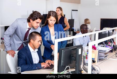 Focused young business people grouped around computer monitor, discussing new project in coworking space Stock Photo