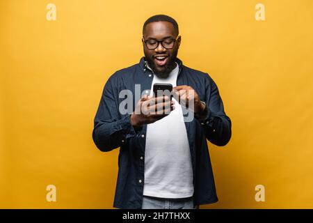 Portrait of cheerful young black guy sending message on mobile phone against yellow background. Happy African-American man using smartphone, enjoying chatting online, messaging in social networks Stock Photo