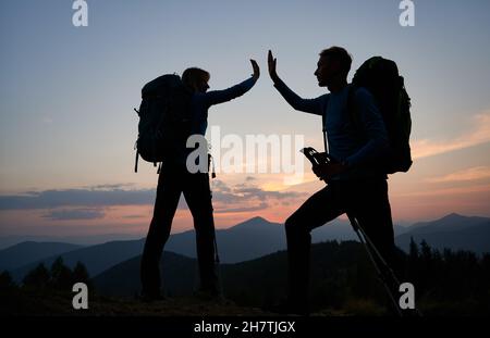 Family pair of hikers fun clapping each other's hands when they reached top of the mountain during outdoor hike. Evening hiking on mountain hills in twilight under evening sky. Concept of travelling. Stock Photo