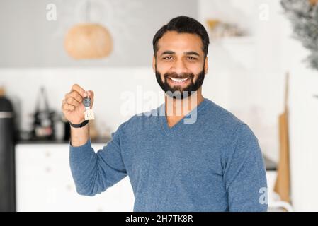 The keys in hand of happy bearded Indian man, cheerful male moved in new house, rent apartment, cheerful ethnic guy bought own real estate, the key with keyring in shape of little house Stock Photo