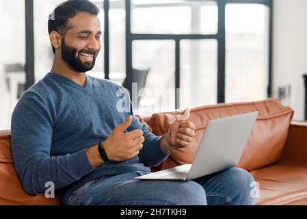 Handsome bearded Indian guy using laptop for video connection, sitting on the sofa at home, smiling ethnic man in casual wear talking online, showing thumbs up into webcam Stock Photo