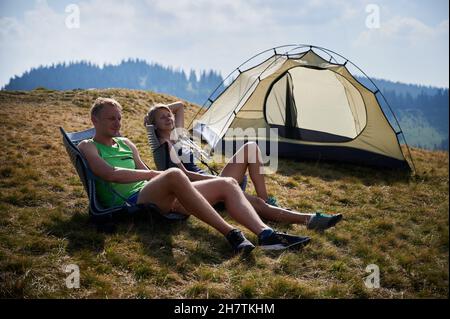 Young woman traveler sitting in tourist chair, relaxing with husband near tourist tent. Happy young couple hikers resting on grassy hill near camp tent while hiking together in mountains. Stock Photo