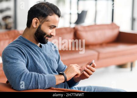 Handsome Indian man sending messages on the smartphone, sitting on the comfortable couch with a cell phone in hands, texting, scrolling and web surfing, side view Stock Photo