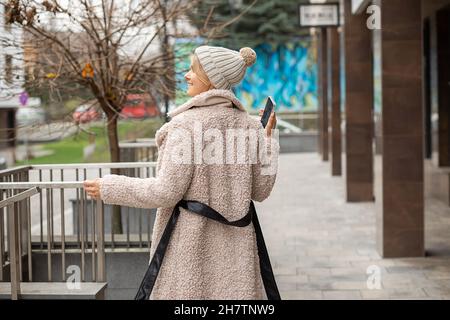 Photo of beautiful young woman with smart phone in hand wearing knitted hat and coat made of faux fur in city among concrete buildings. Happy life, re Stock Photo