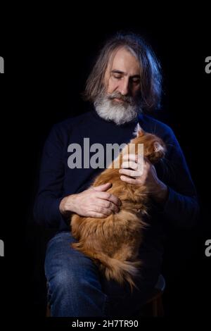 Elderly owned and ginger cat. Portrait of an elderly gray-haired bearded man in dark clothes on a dark background. Stock Photo