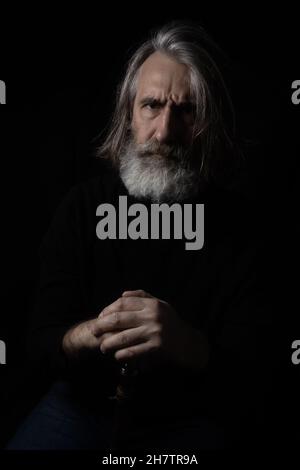 Portrait of an elderly gray-haired bearded man in dark clothes on a dark background. The concept is aging, sadness, wisdom. Stock Photo