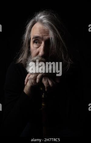 An elderly gray-haired bearded man rests his hands and chin on the knob of an old cane Stock Photo