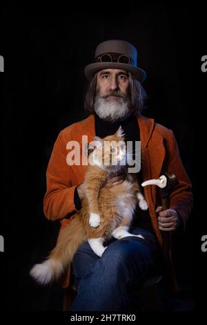 A gray-haired bearded intellectual of mature age in a steampunk hat poses with a ginger cat. Stock Photo