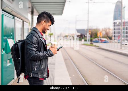stylish latin student with coffee in hand consults his phone while waiting for public transportation to arrive at the streetcar stop, concept of susta Stock Photo