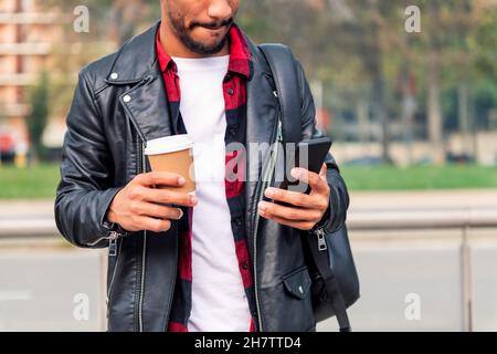 detail of the hands of a stylish young latin student consulting his phone with a take away coffee, concept of technology and urban lifestyle Stock Photo
