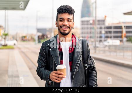 stylish young latin guy smiles happily looking at camera with a take away coffee in his hand, concept of urban lifestyle Stock Photo
