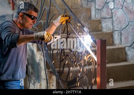Man wearing protective glasses welds metal with welding machine. Sparks from welding fly to sides. Stock Photo