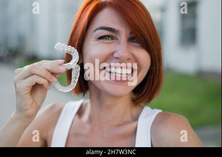 Red-haired Caucasian woman holding transparent mouthguards for bite correction outdoors. A girl with a beautiful snow-white smile uses silicone braces Stock Photo