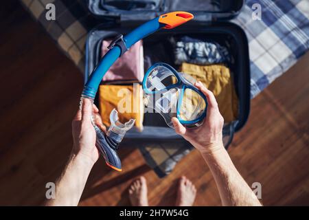 Young man holding snorkel and goggles in hands at home. Packing suitcase and planning travel adventure. Stock Photo