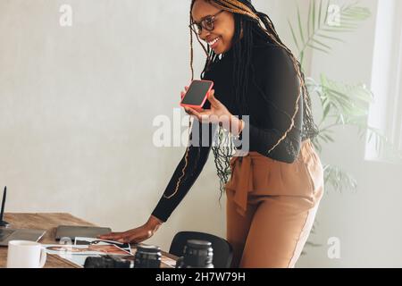 Creative freelancer taking a phone call in her home office. Happy young woman smiling while standing at a desk with photographic equipment. Female pho Stock Photo